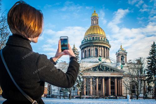 Исаакиевский собор в Санкт-Петербурге — жемчужина архитектуры в зимнем великолепии. На заднем плане — музеи и архитектурные богатства города. Туристка, запечатленная на фотографии, делает снимки на своем телефоне, зафиксировав момент путешествия в Россию и красоты Петербурга.